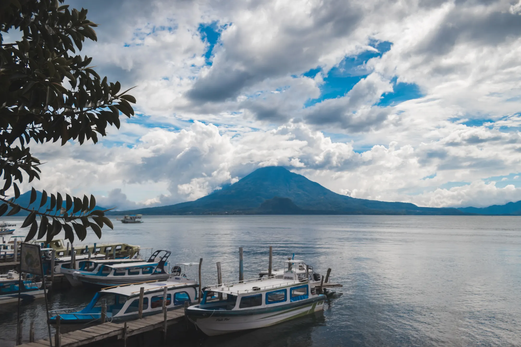 Boats docked on Lake Atitlán beneath a towering volcano and dramatic clouds